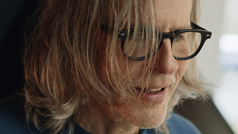 [Photo] Close-up of a person wearing glasses, with shoulder-length hair partially covering the face, photographed in soft indoor light.