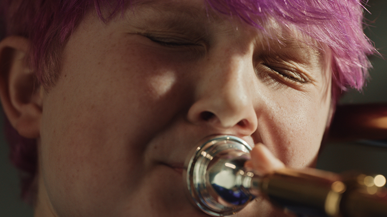 [Photo] Close-up of a person playing a brass instrument, with the mouthpiece pressed to the lips and the face tightly framed.