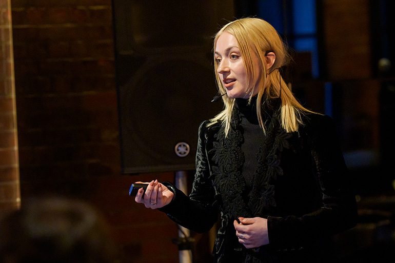[Photo] A female presenter delivering a presentation on stage with expressive gestures during the TRANSPOSE Innovation Challenge final pitch.