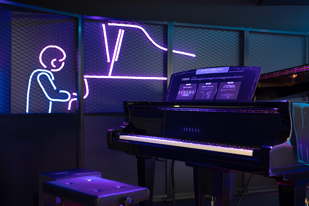 [ 画像 ] ピアノを近くから見た画像。すぐそばに、ピアノを弾いている人をかたどったネオンライトがある。