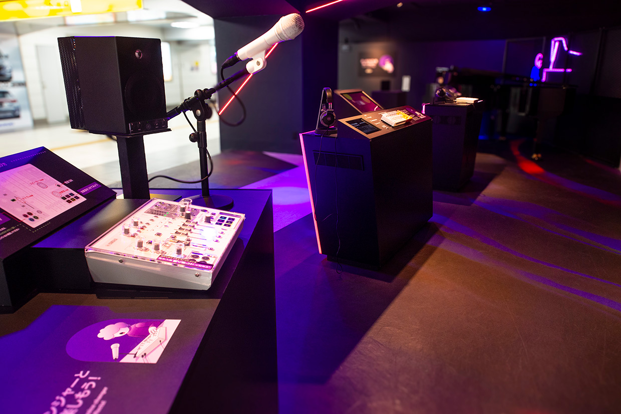 [ 画像 ] 展示ブースの中。手前の3つの台の上に小型の製品があり、奥にはピアノが設置されている。