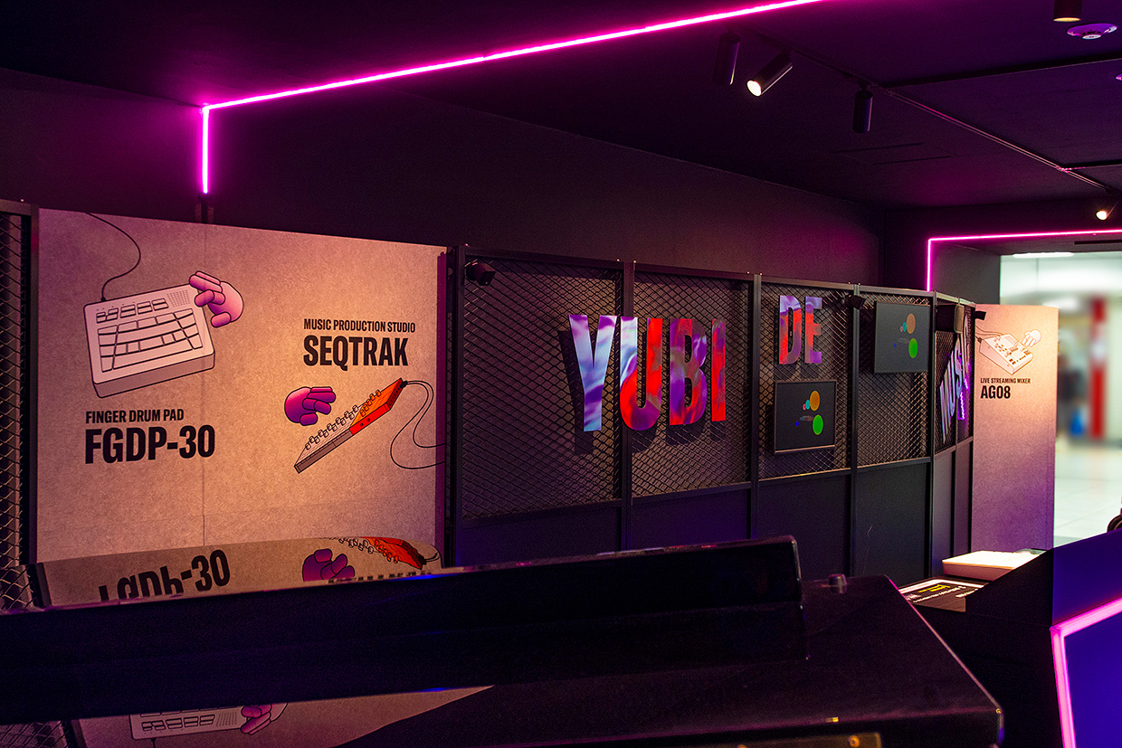 [ 画像 ] 展示ブースの中。壁やフェンスに製品のイラストや文字があしらわれている。