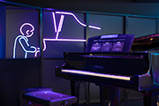 [ 画像 ] ピアノを近くから見た画像。すぐそばに、ピアノを弾いている人をかたどったネオンライトがある。