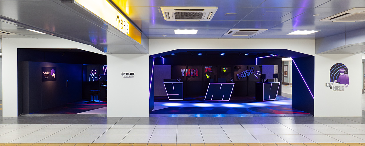 展示ブースイメージ。床は赤や紫が混ざり合う柄で、グレーの壁に黒いフェンスやネオンサインを設置。楽器や音響機器が展示されている。