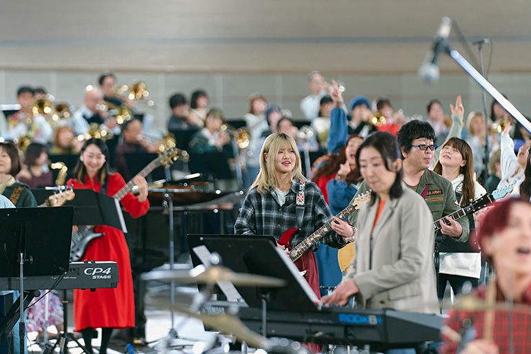 [写真] キーボードやギター、管楽器など多様な楽器の参加者が大人数で合奏している様子