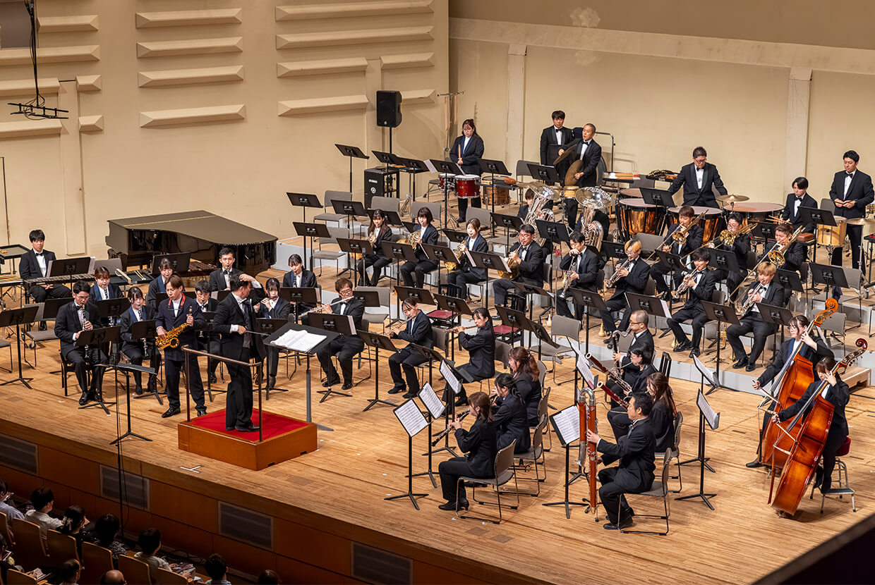 [サムネイル] コンサートホールで演奏中の大規模な吹奏楽団。指揮者を中心に木管楽器、金管楽器、打楽器、弦楽器が配置され、観客席も一部見える。