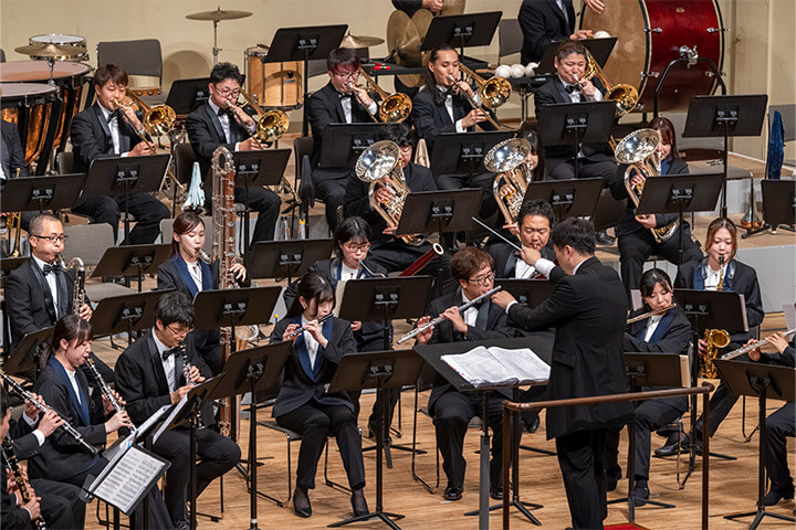 [写真] オーケストラの演奏風景、指揮者が前に立ち管楽器奏者が多数並んでいる。演奏者はフォーマルな服装で、トランペットやフルートなどさまざまな楽器を演奏している様子が見られる。