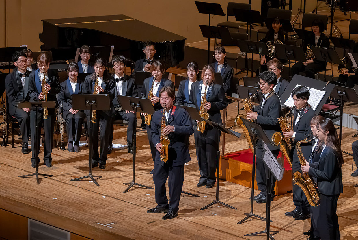 [写真] 演奏会のステージでサクソフォン奏者の上野耕平さんが立って演奏している。複数の演奏者が並び、後方にはピアノや他の楽器奏者が配置されている。