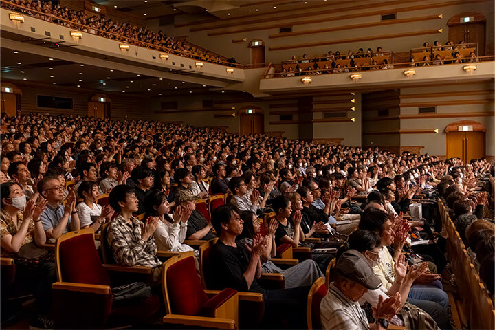 [写真] ホール内で多くの観客が舞台に向かって拍手している。観客席は複数の階層に分かれ、温かみのある照明と木目調の内装が特徴的。