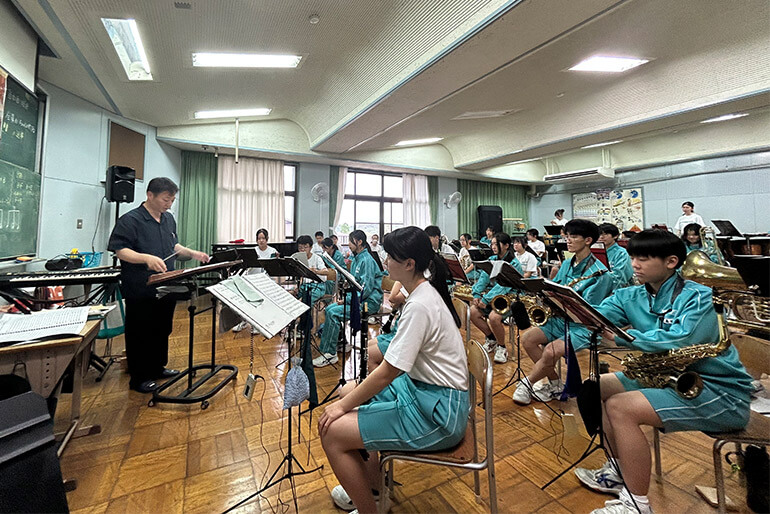 [写真] 吹奏楽部の練習風景、学生たちが楽器を演奏し指導者が指示を出している様子が見られる。教室内で楽譜スタンドや楽器が整然と配置され、集中して演奏に取り組む姿が特徴的。