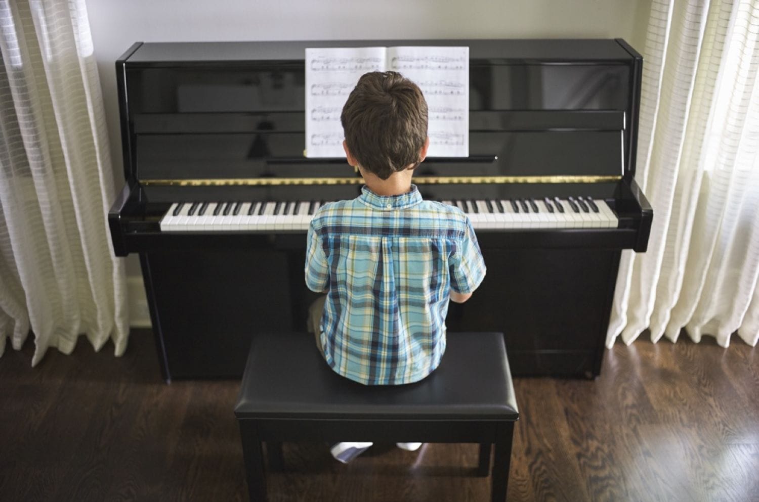 Small boy with back to camera as he is sitting at piano playing while looking at sheet music on ledge.