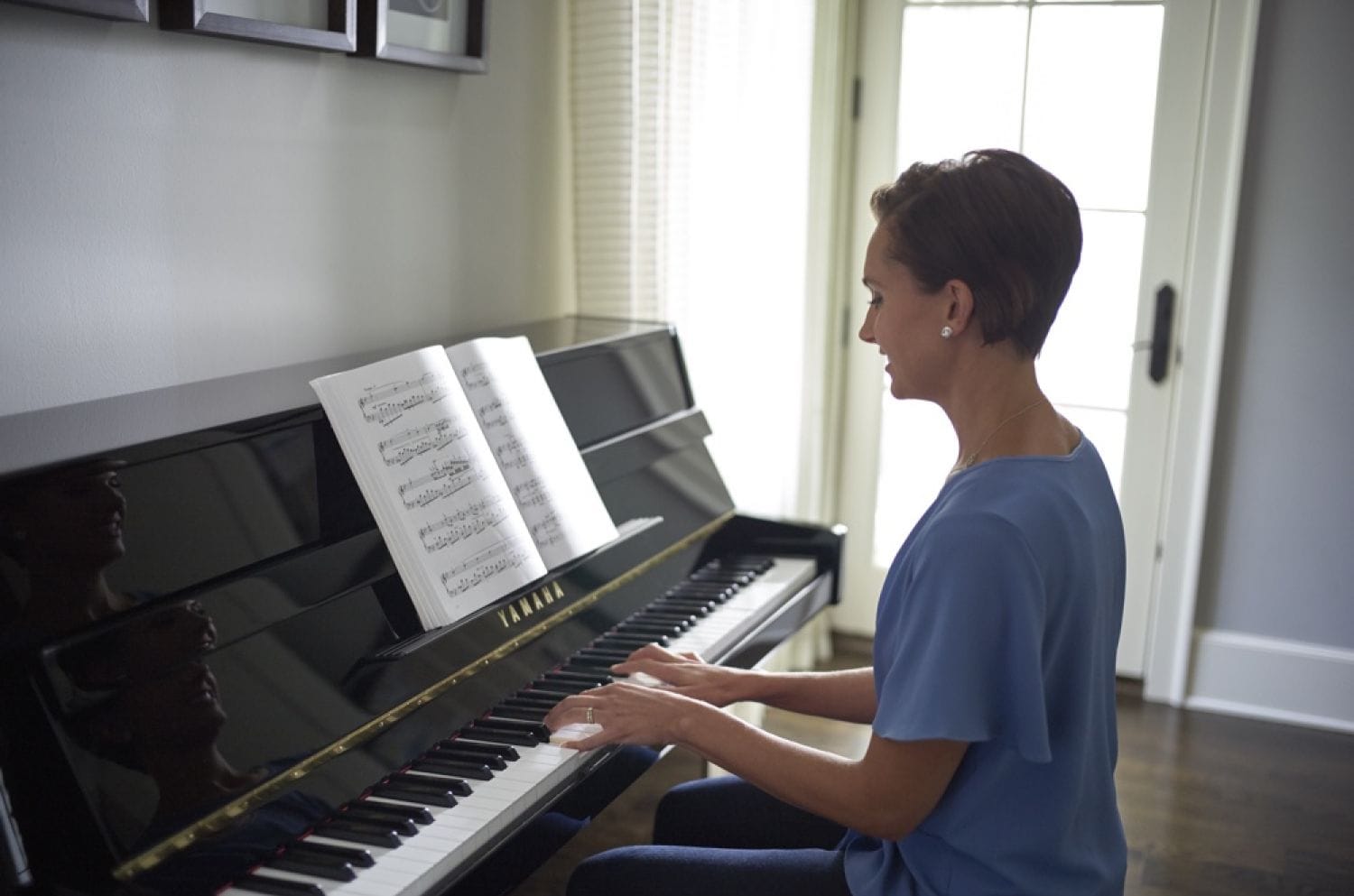 Woman in her early 30's in her living room playing an upright piano from book of sheet music.