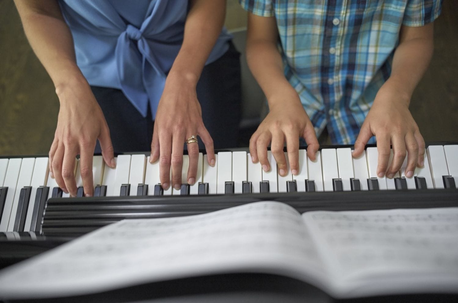 Two pairs of hands on keys viewed from above. You can see a woman's hands with wedding rings and a small child's hands. There is an open book of music on ledge above keys.