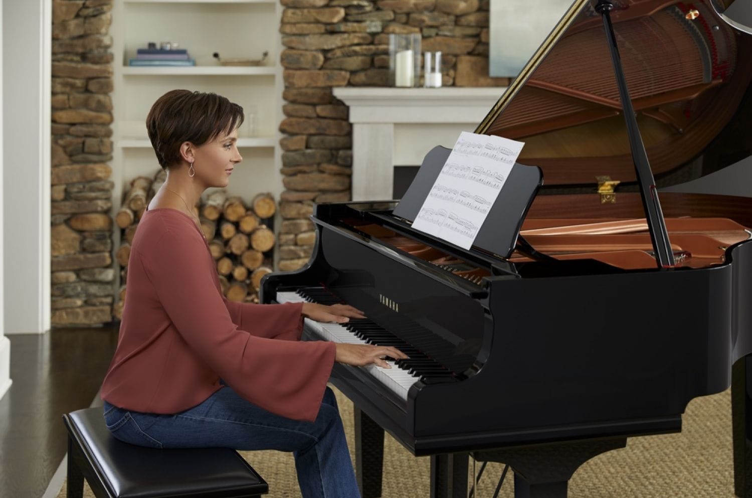 Young woman in casual clothes playing Yamaha baby grand from sheet music with fireplace and bookshelves in background.