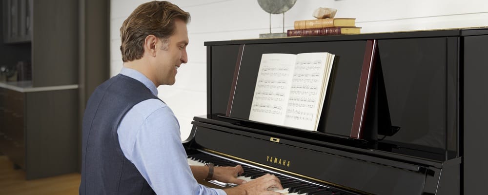 A man smiling as he plays from sheet music on a Yamaha upright piano in his living room.