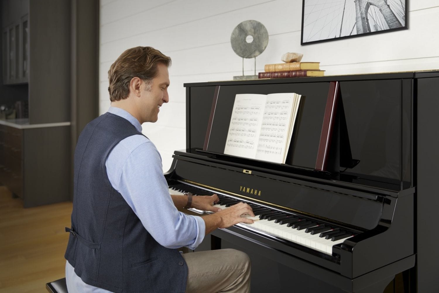 A man approximately 40 years old is smiling broadly while playing from sheet music on a Yamaha upright piano in a living room setting.