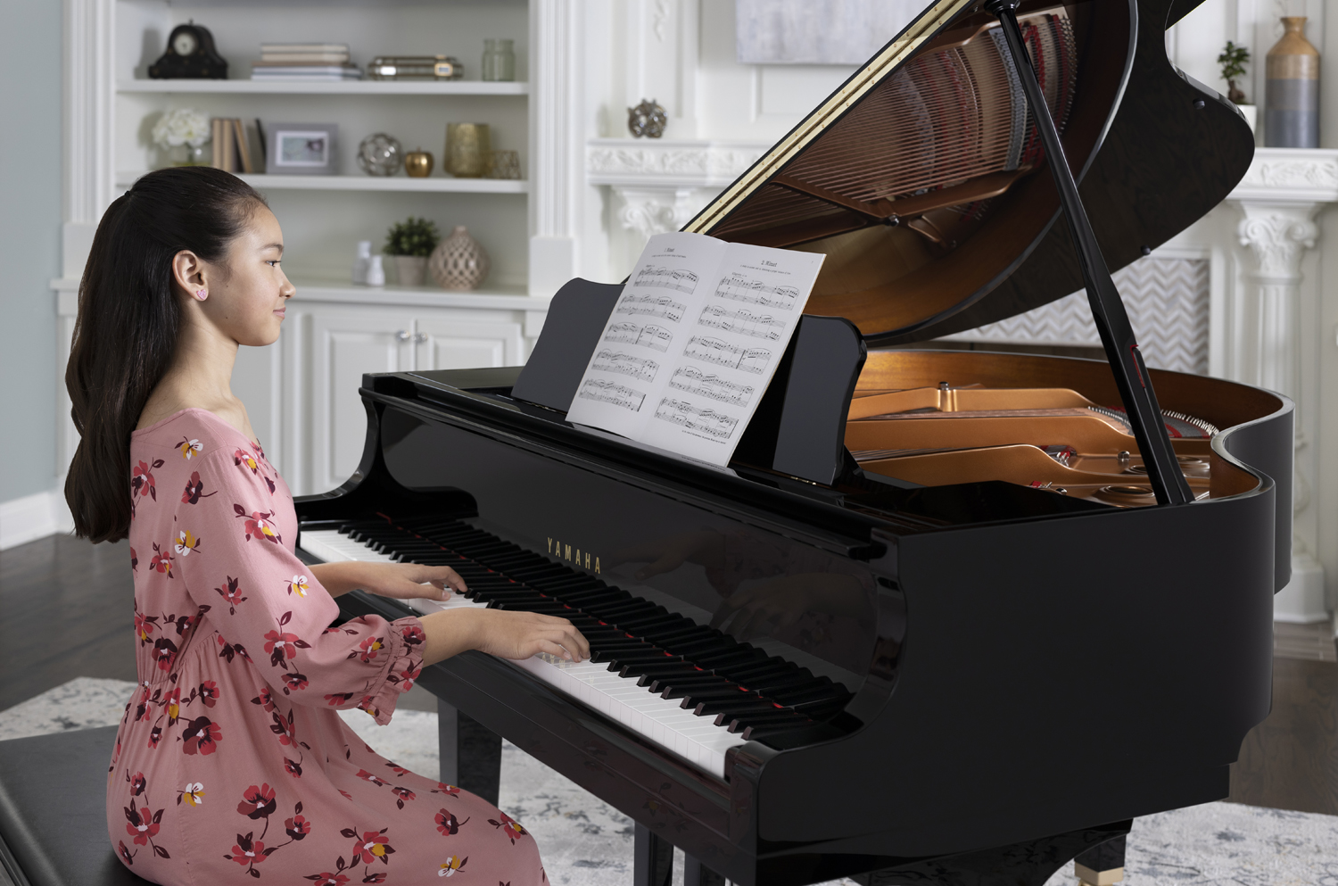 Young woman in casual clothes playing Yamaha baby grand from sheet music with fireplace and bookshelves in background.