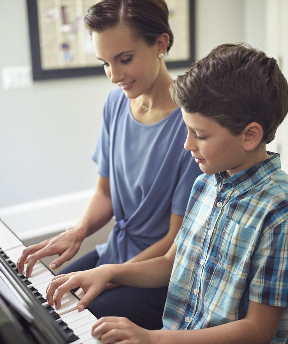 A woman in her 30's next to her young son at the upright piano showing him how to play with his right hand. He's imitating her.