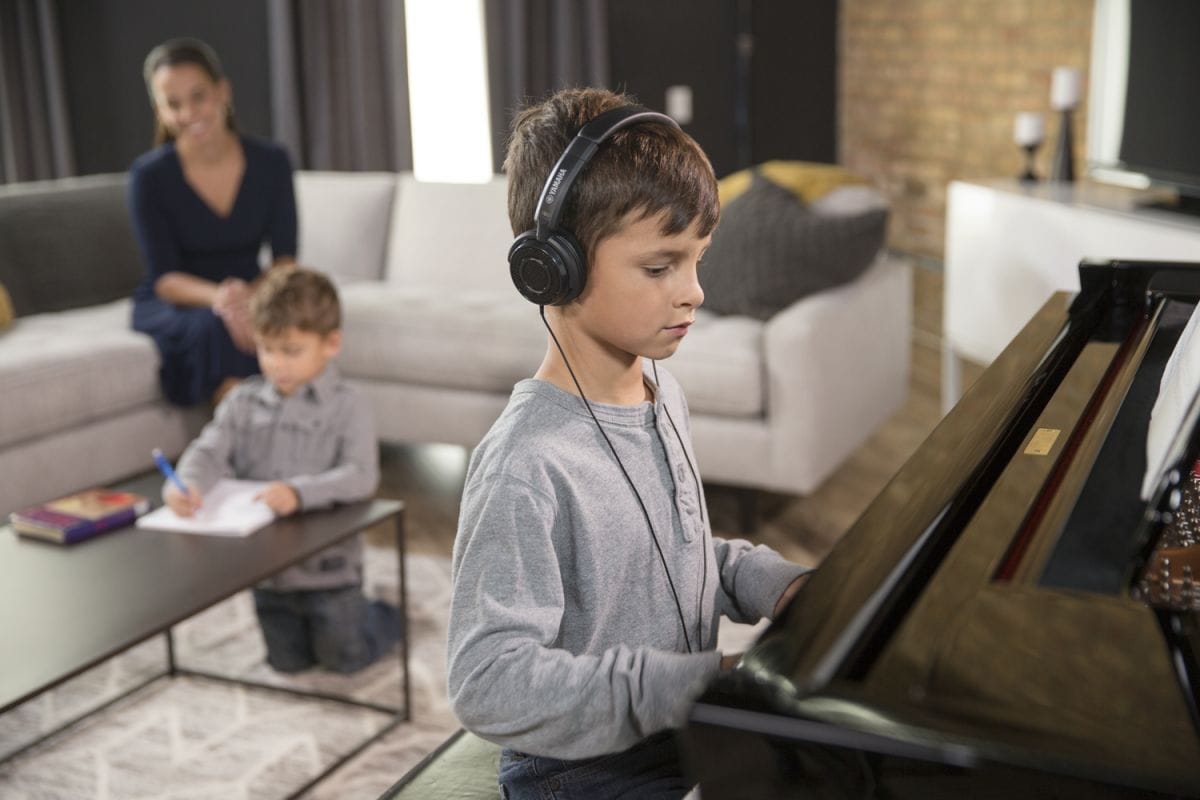 Young boy playing an upright piano in his living room wearing headphones while his mother and younger brother hang out undisturbed behind him. 